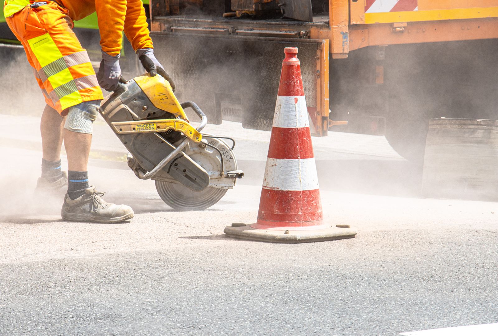 Construction worker cutting asphalt with a saw, creating dust on a hot day